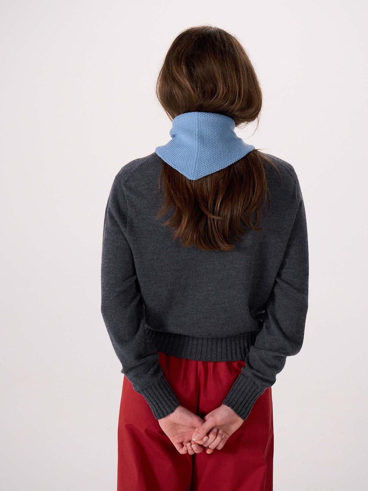 A person stands with their back to the camera, wearing a dark gray sweater, red pants, and the Daisy Scarf – Celeste Blue draped around their neck. Their hands are clasped behind their back. The scarf measures 22cm wide and 97cm long.
