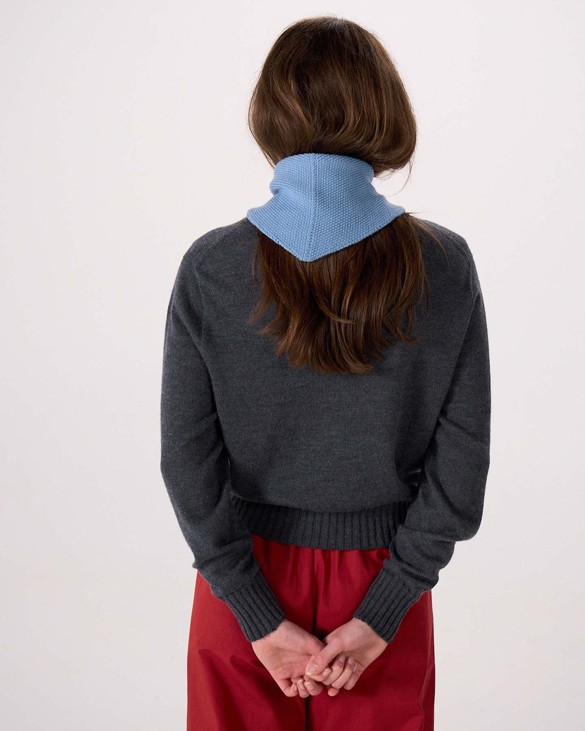 A person stands with their back to the camera, wearing a dark gray sweater, red pants, and the Daisy Scarf – Celeste Blue draped around their neck. Their hands are clasped behind their back. The scarf measures 22cm wide and 97cm long.