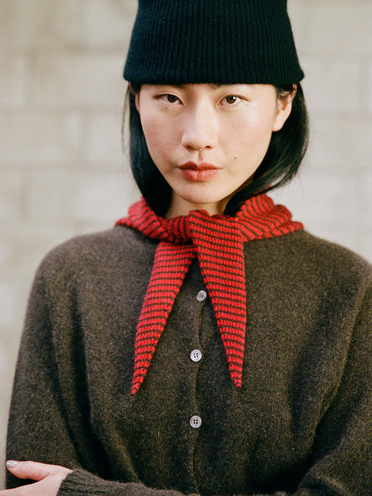 A person in a black beanie, brown buttoned sweater, and the 97cm Daisy Scarf – Cherry & Chestnut Stripe tied in front, stands against a light brick wall.