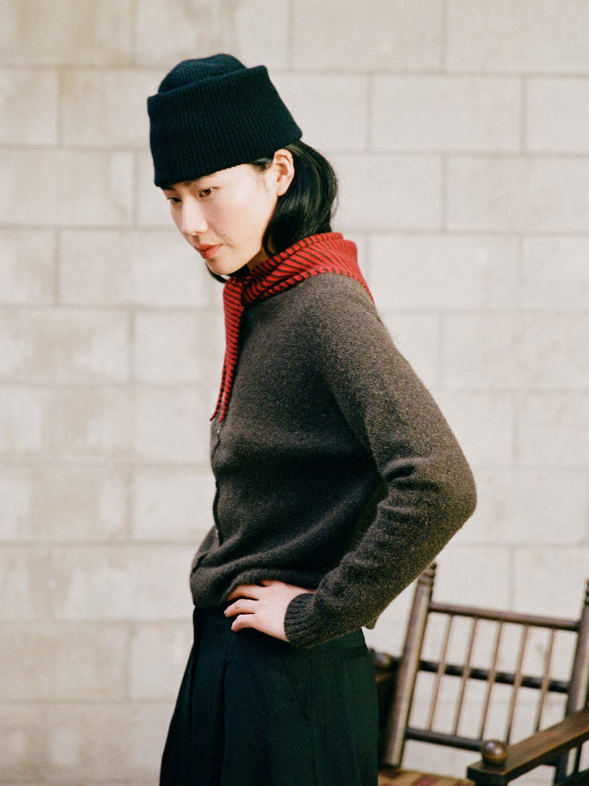 A person wearing a dark sweater, black beanie, and the Daisy Scarf – Cherry & Chestnut Stripe (97cm x 22cm) stands sideways with one hand on their hip in front of a light brick wall and wooden chair.