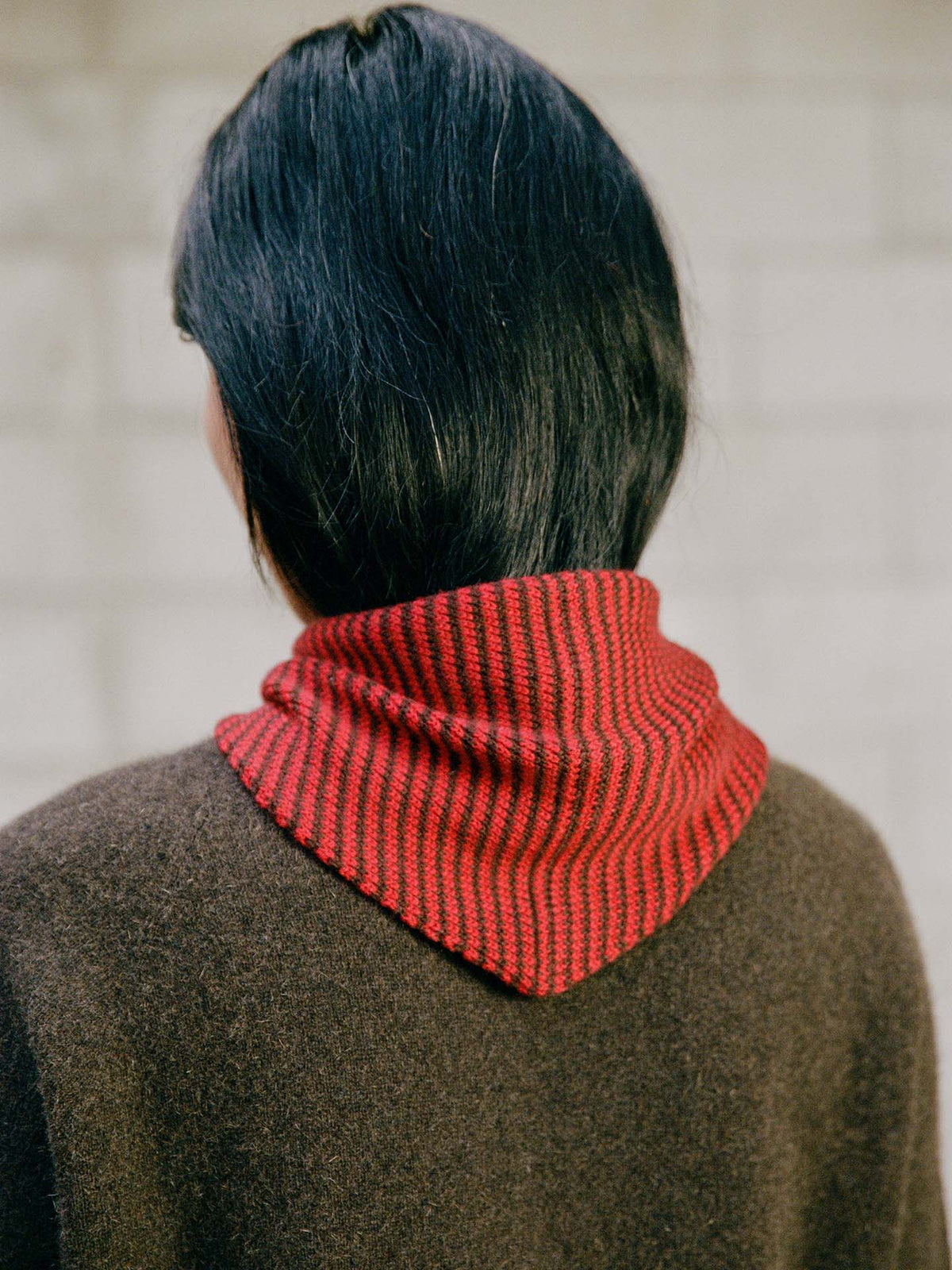 Person with straight, black shoulder-length hair shown from the back, wearing a brown top and the Daisy Scarf – Cherry & Chestnut Stripe (97cm long x 22cm wide), a red-and-black striped knit neck scarf.