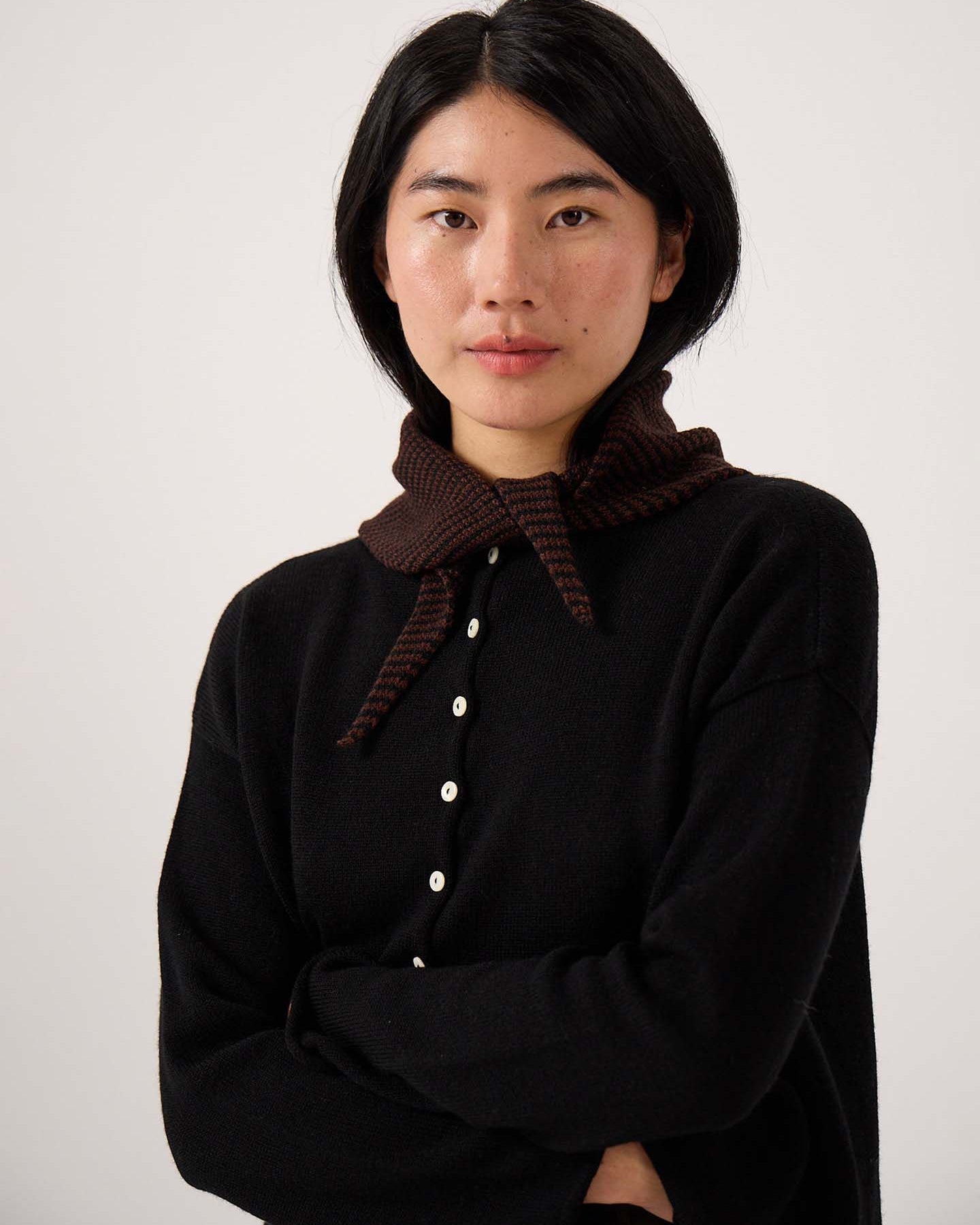 A woman with straight black hair and a neutral expression wears a black button-up top accessorized with the Daisy Scarf – Chestnut & Black Stripe, standing against a plain light background with her arms crossed. The scarf measures 97 cm in length and 22 cm in width.