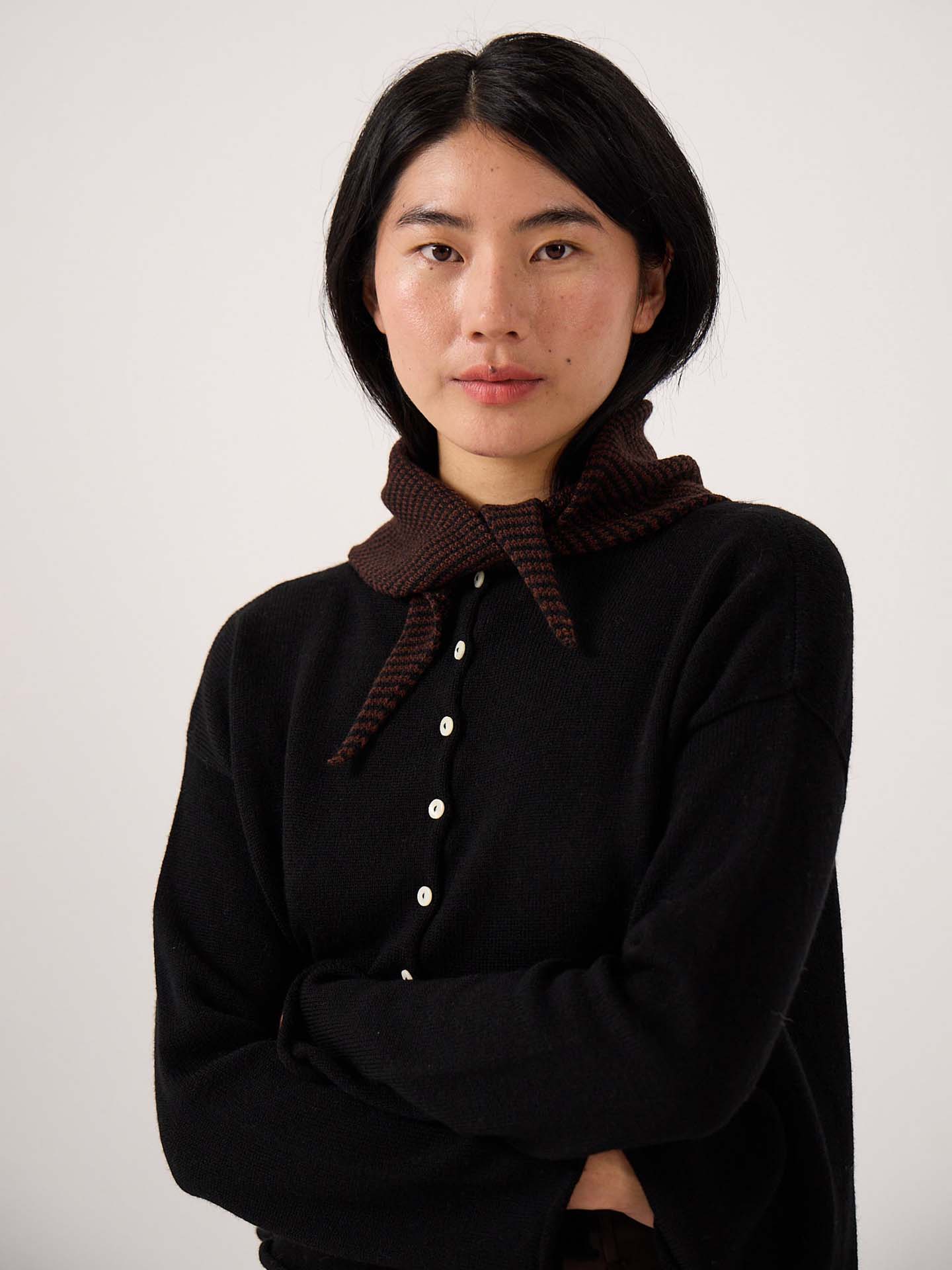 A woman with straight black hair and a neutral expression wears a black button-up top accessorized with the Daisy Scarf – Chestnut & Black Stripe, standing against a plain light background with her arms crossed. The scarf measures 97 cm in length and 22 cm in width.
