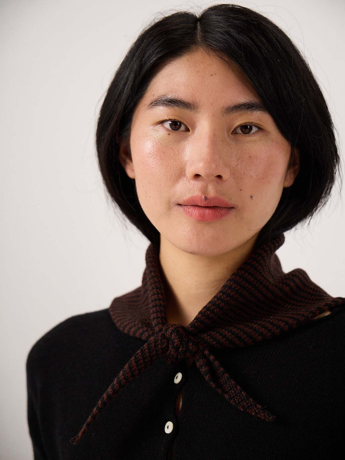 A person with short black hair, dressed in a black top with white buttons and wearing the Daisy Scarf – Chestnut & Black Stripe—a dark brown knitted scarf tied at the neck and measuring 22cm wide by 97cm long—is looking directly at the camera.