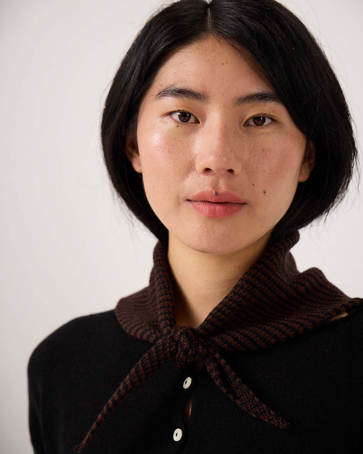 A person with short black hair, dressed in a black top with white buttons and wearing the Daisy Scarf – Chestnut & Black Stripe—a dark brown knitted scarf tied at the neck and measuring 22cm wide by 97cm long—is looking directly at the camera.