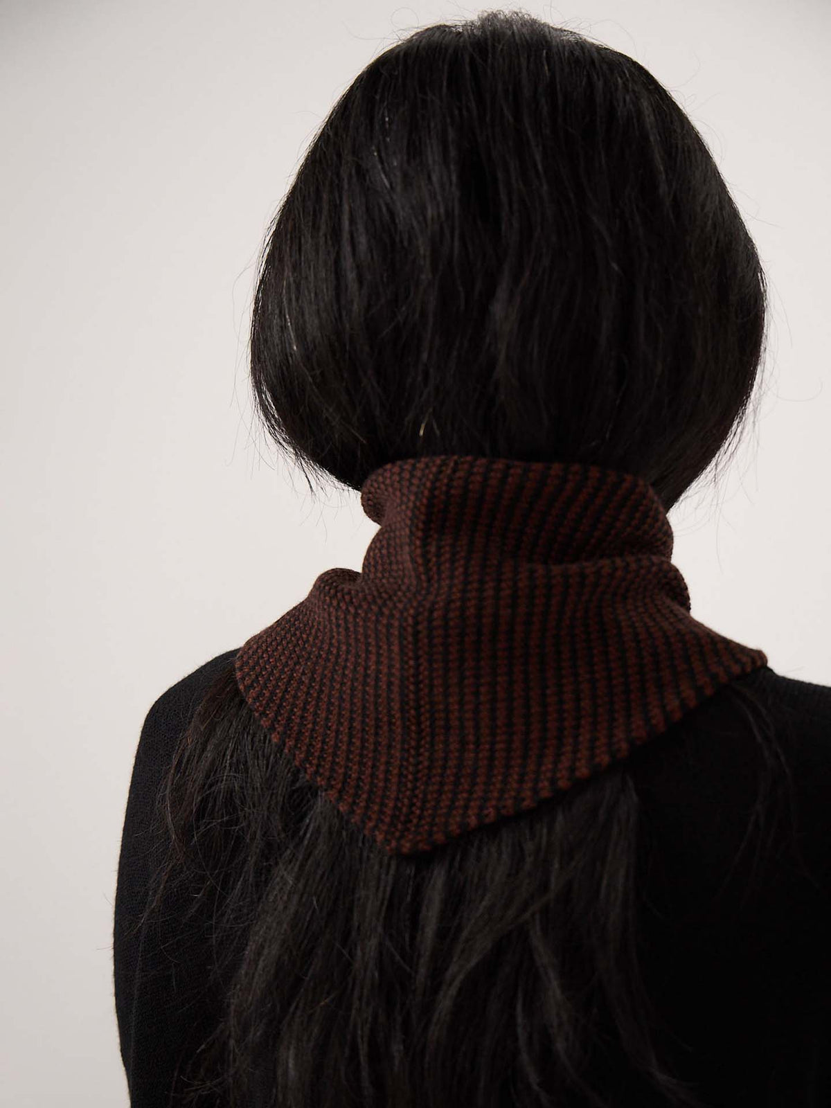 Person with long dark hair facing away, wearing a black top and the Daisy Scarf – Chestnut & Black Stripe, a dark brown knitted scarf with black stripes measuring 97cm long and 22cm wide, wrapped around their neck against a plain background.