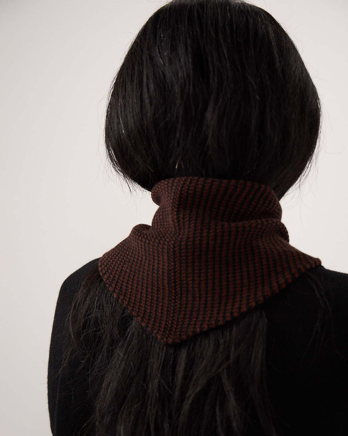Person with long dark hair facing away, wearing a black top and the Daisy Scarf – Chestnut & Black Stripe, a dark brown knitted scarf with black stripes measuring 97cm long and 22cm wide, wrapped around their neck against a plain background.
