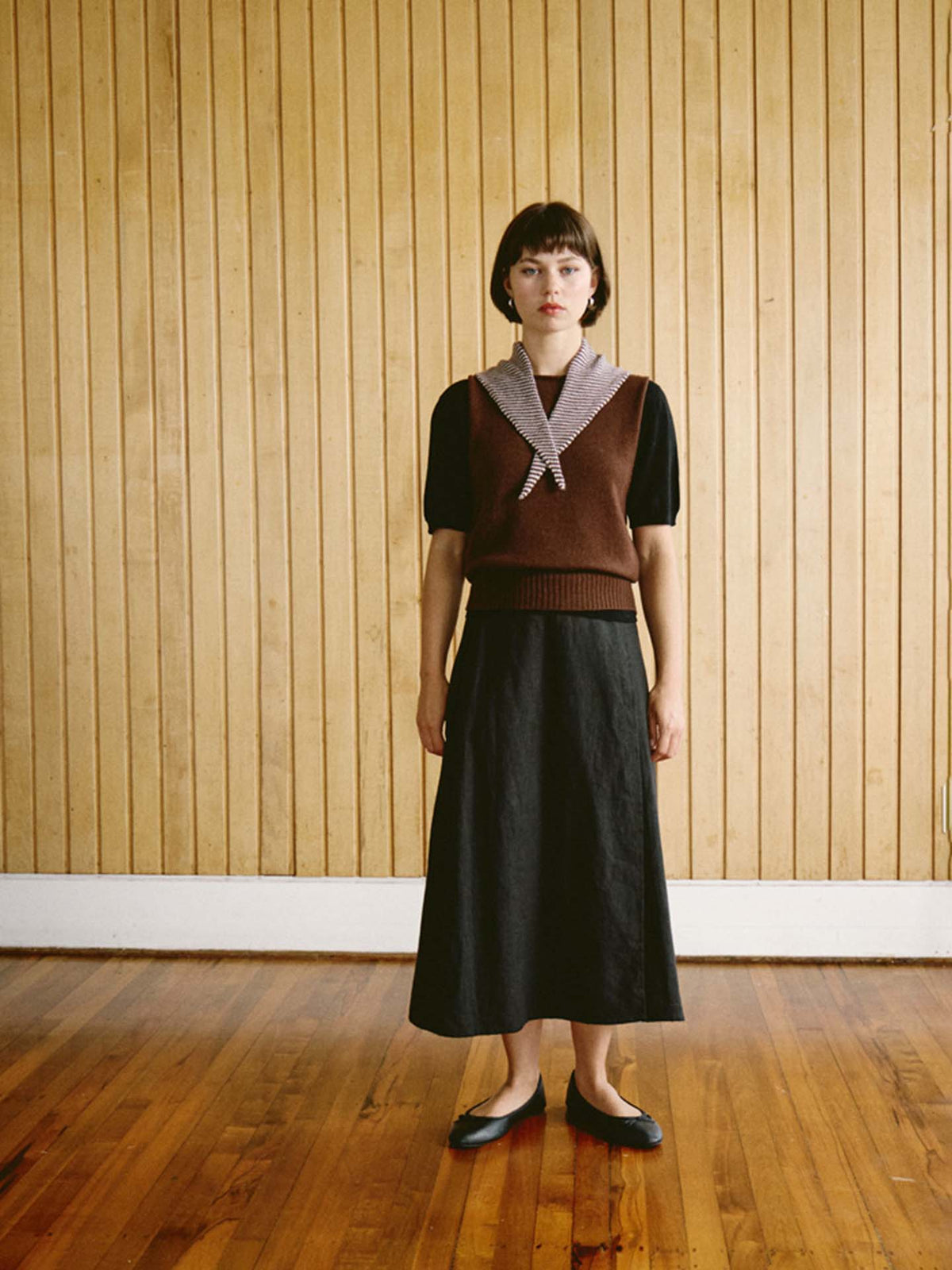 A woman stands indoors on a wooden floor in front of a vertical wood-paneled wall, wearing a brown sweater vest, black skirt (97 cm long and 22 cm wide), black shoes, and the Daisy Scarf – Chestnut & Ivory Stripe. The outfit has a sleek and tailored appearance.