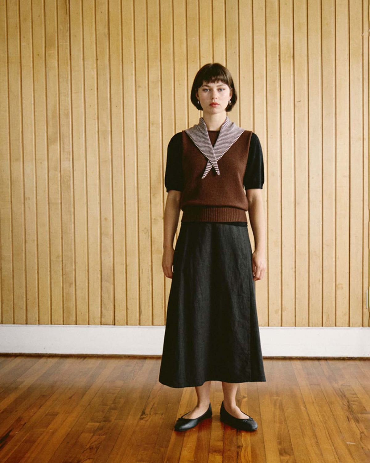 A woman stands indoors on a wooden floor in front of a vertical wood-paneled wall, wearing a brown sweater vest, black skirt (97 cm long and 22 cm wide), black shoes, and the Daisy Scarf – Chestnut & Ivory Stripe. The outfit has a sleek and tailored appearance.