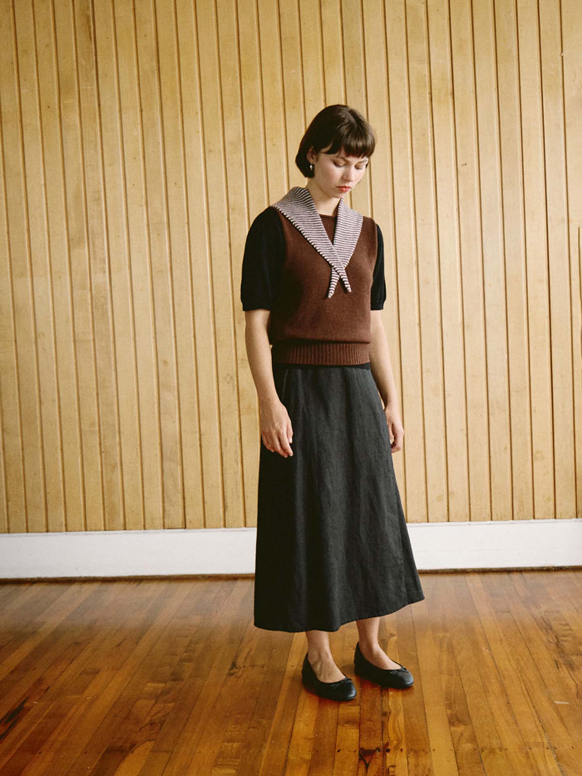 A woman stands indoors on a wooden floor in front of a vertical wood panel wall, wearing a brown vest, black shirt, black skirt with refined proportions (97cm long and 22cm wide at the hem), flat shoes, and the Daisy Scarf – Chestnut & Ivory Stripe, adding a touch of sophistication to her stylish ensemble.
