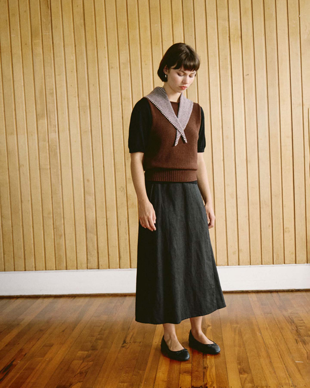 A woman stands indoors on a wooden floor in front of a vertical wood panel wall, wearing a brown vest, black shirt, black skirt with refined proportions (97cm long and 22cm wide at the hem), flat shoes, and the Daisy Scarf – Chestnut & Ivory Stripe, adding a touch of sophistication to her stylish ensemble.