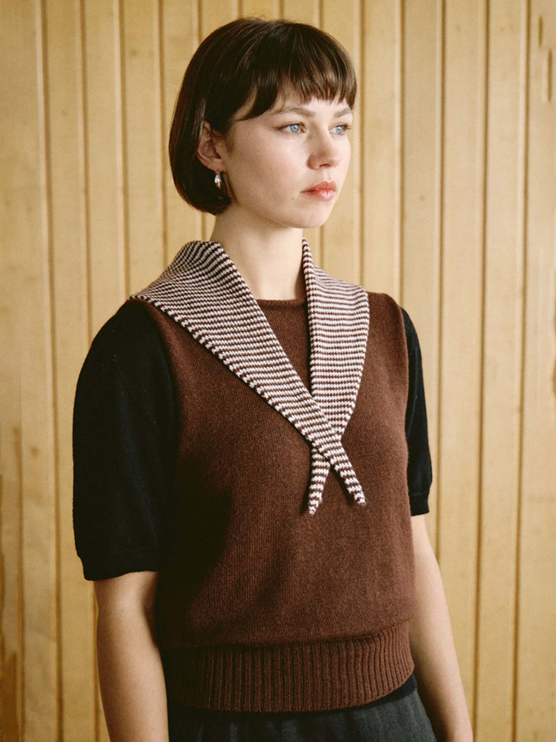 A woman with short brown hair wears a brown sleeveless sweater over a black top, with the Daisy Scarf – Chestnut & Ivory Stripe (measuring 97cm long and 22cm wide) draped around her neck, standing in front of a wooden panel wall.