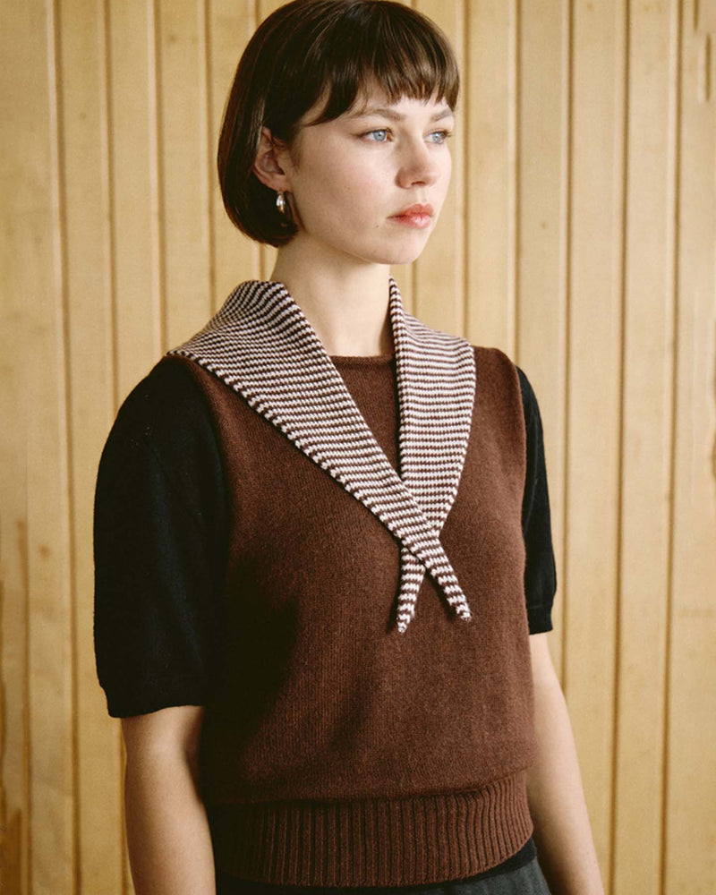 A woman with short brown hair wears a brown sleeveless sweater over a black top, with the Daisy Scarf – Chestnut & Ivory Stripe (measuring 97cm long and 22cm wide) draped around her neck, standing in front of a wooden panel wall.