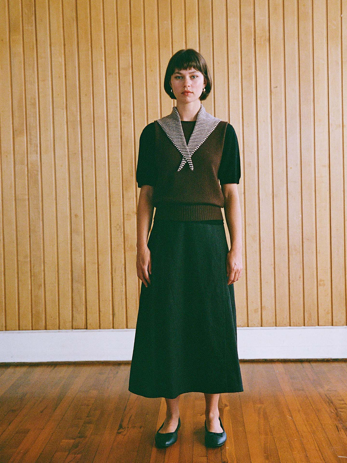 A person with straight brown hair stands indoors against a wooden wall, wearing the Daisy Scarf – Chestnut & Ivory Stripe, perfectly showcasing its elegant design.
