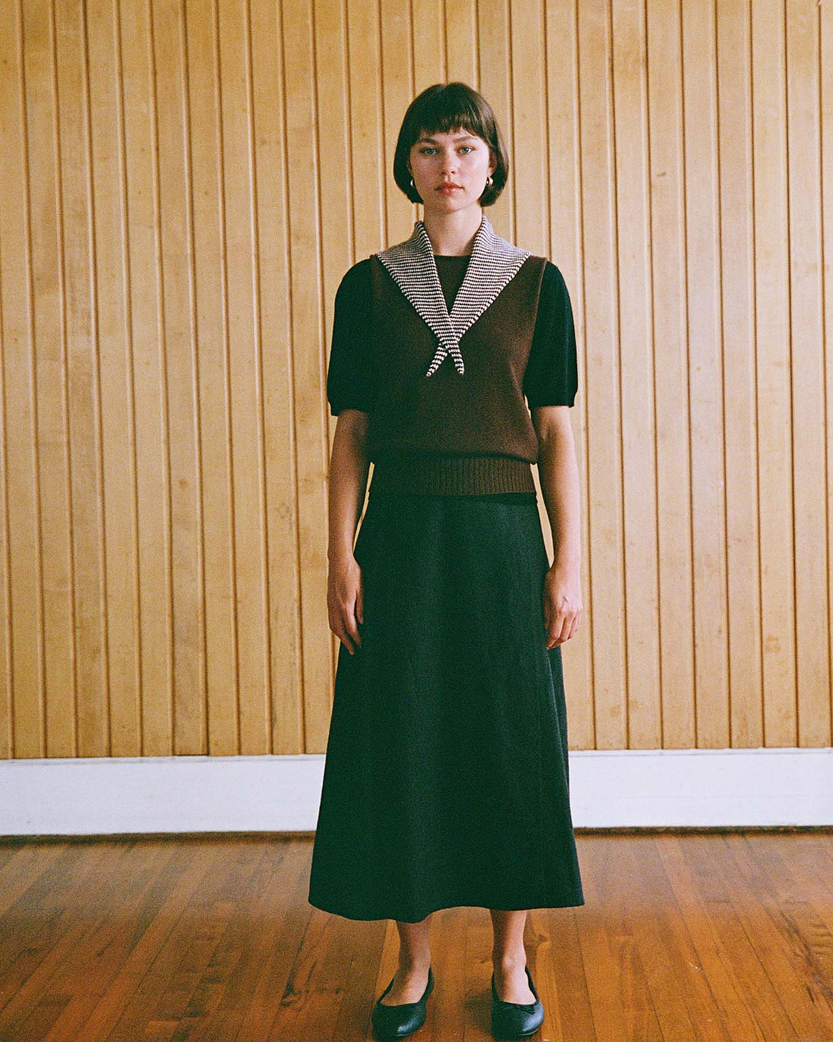 A person with straight brown hair stands indoors against a wooden wall, wearing the Daisy Scarf – Chestnut & Ivory Stripe, perfectly showcasing its elegant design.