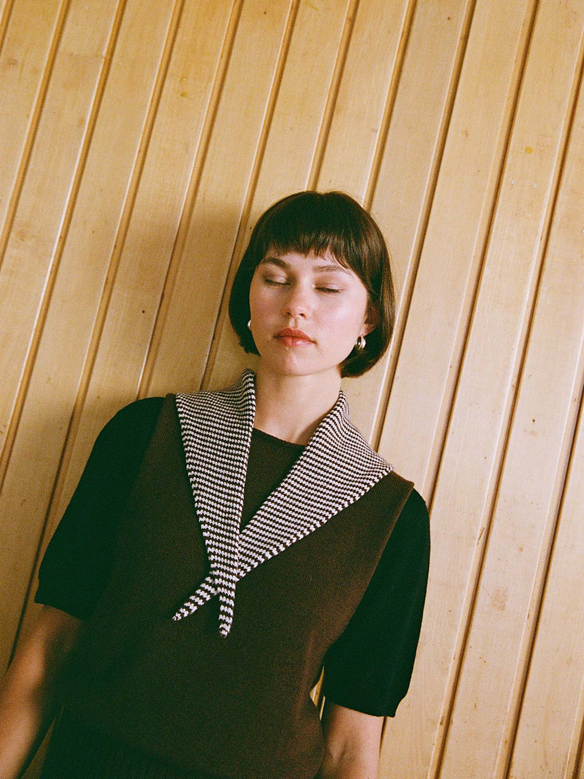 A person with short brown hair and closed eyes stands against a vertical wooden panel wall, wearing a black top accented by the Daisy Scarf – Chestnut & Ivory Stripe. The wooden panels, each 22cm wide and neatly aligned along the wall, create a textured backdrop that enhances the tranquil atmosphere.