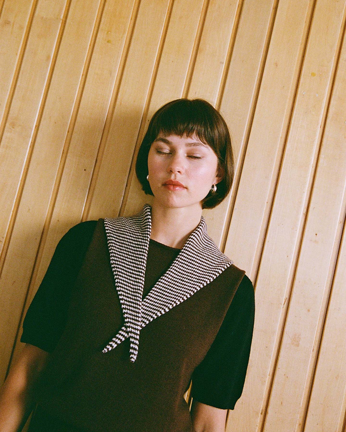 A woman with short brown hair wears a brown sleeveless sweater over a black top, with the Daisy Scarf – Chestnut & Ivory Stripe (measuring 97cm long and 22cm wide) draped around her neck, standing in front of a wooden panel wall.