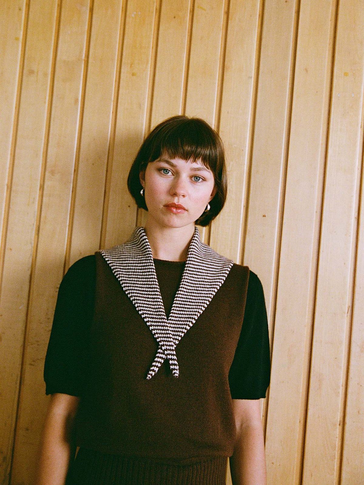 A woman with short brown hair stands in front of a wooden wall, wearing a brown top and the Daisy Scarf – Chestnut & Ivory Stripe draped around her neck. Measuring 97cm long and 22cm wide, the scarf adds an elegant touch to her look while highlighting its stylish dimensions.