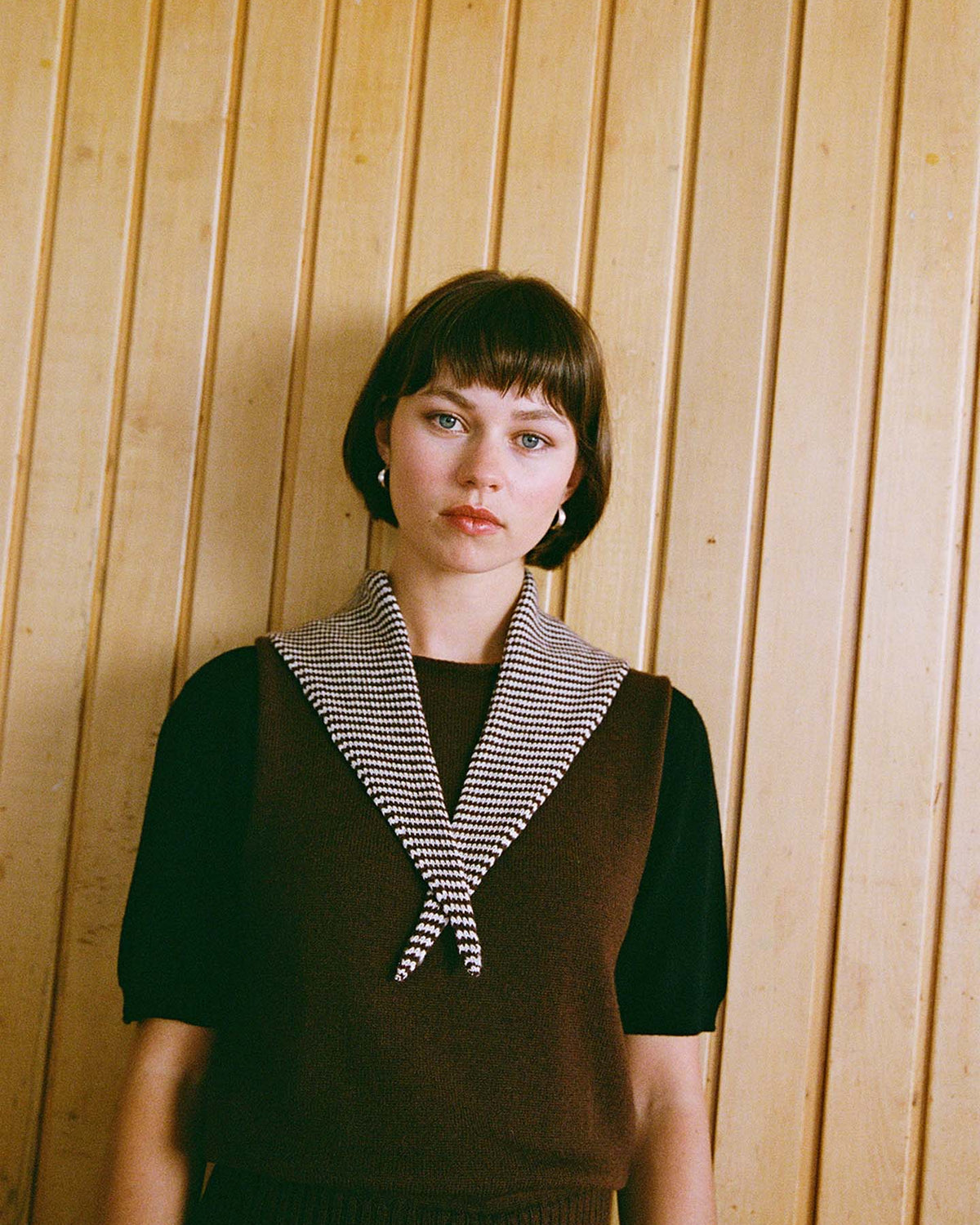 A woman with short brown hair stands in front of a wooden wall, wearing a brown top and the Daisy Scarf – Chestnut & Ivory Stripe draped around her neck. Measuring 97cm long and 22cm wide, the scarf adds an elegant touch to her look while highlighting its stylish dimensions.