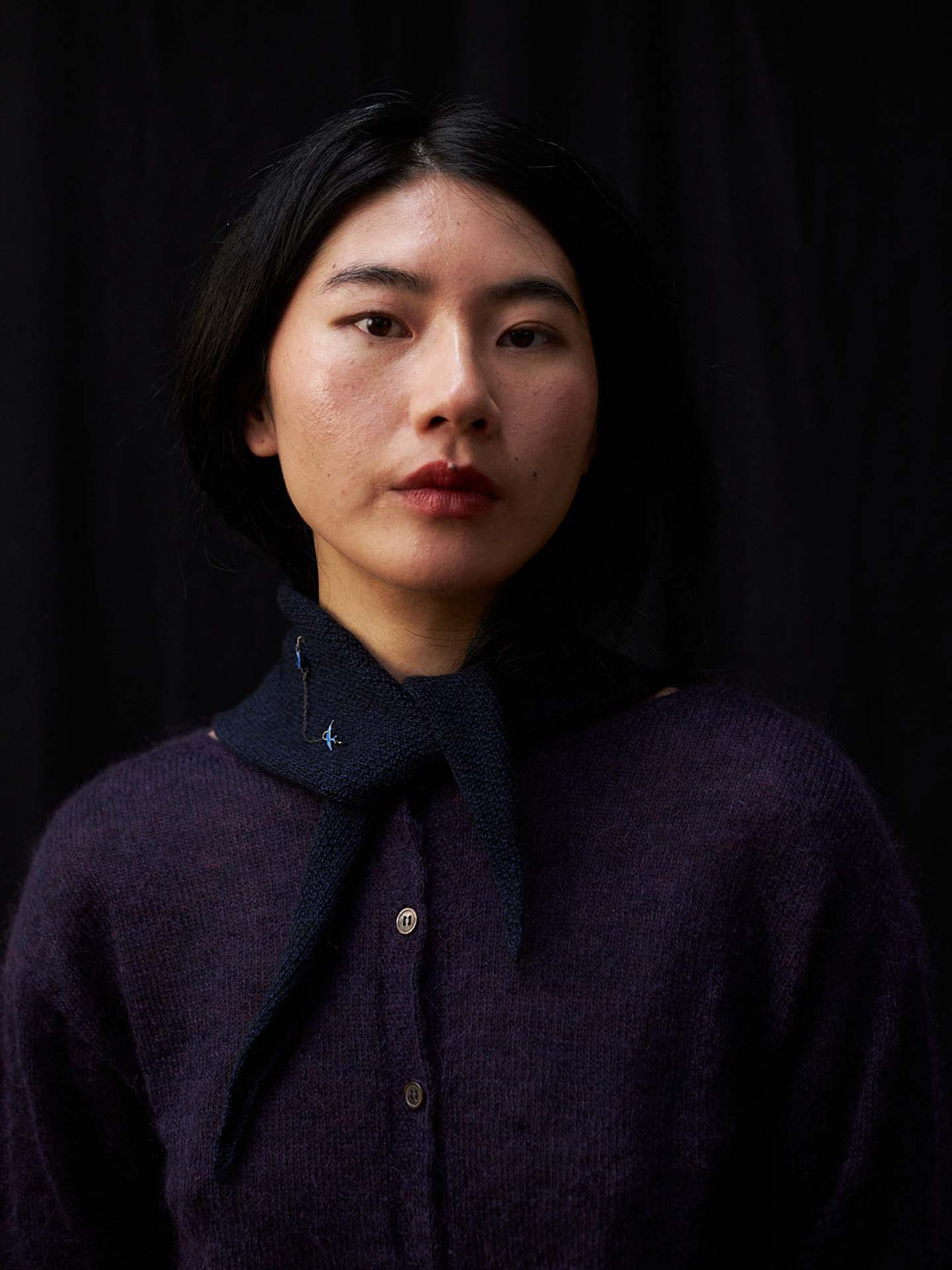 A person with straight black hair wears a dark purple sweater and the Daisy Scarf – Ink (22cm x 97cm), posing in front of a black background. The scarf adds a refined touch to the look.