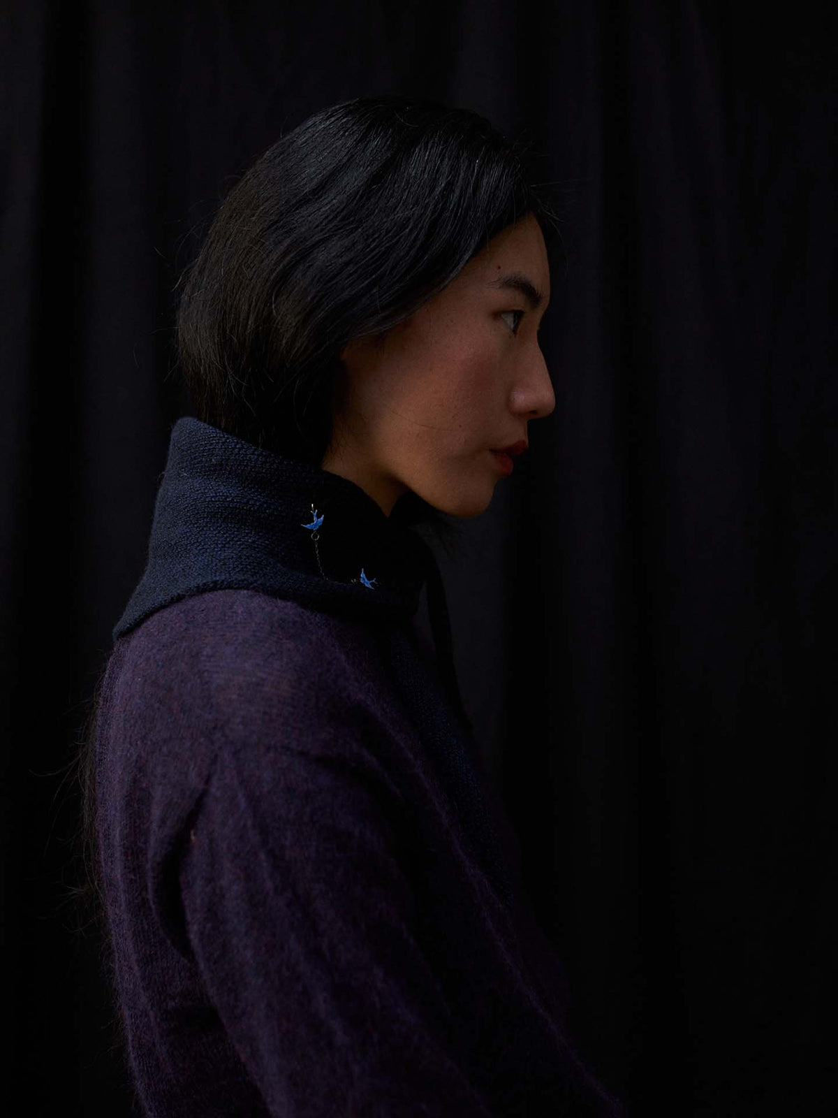 A person with long dark hair, wearing a dark purple sweater and the Daisy Scarf – Ink (97cm long, 22cm wide), is shown in profile against a black background, highlighting the scarf’s elegant silhouette.