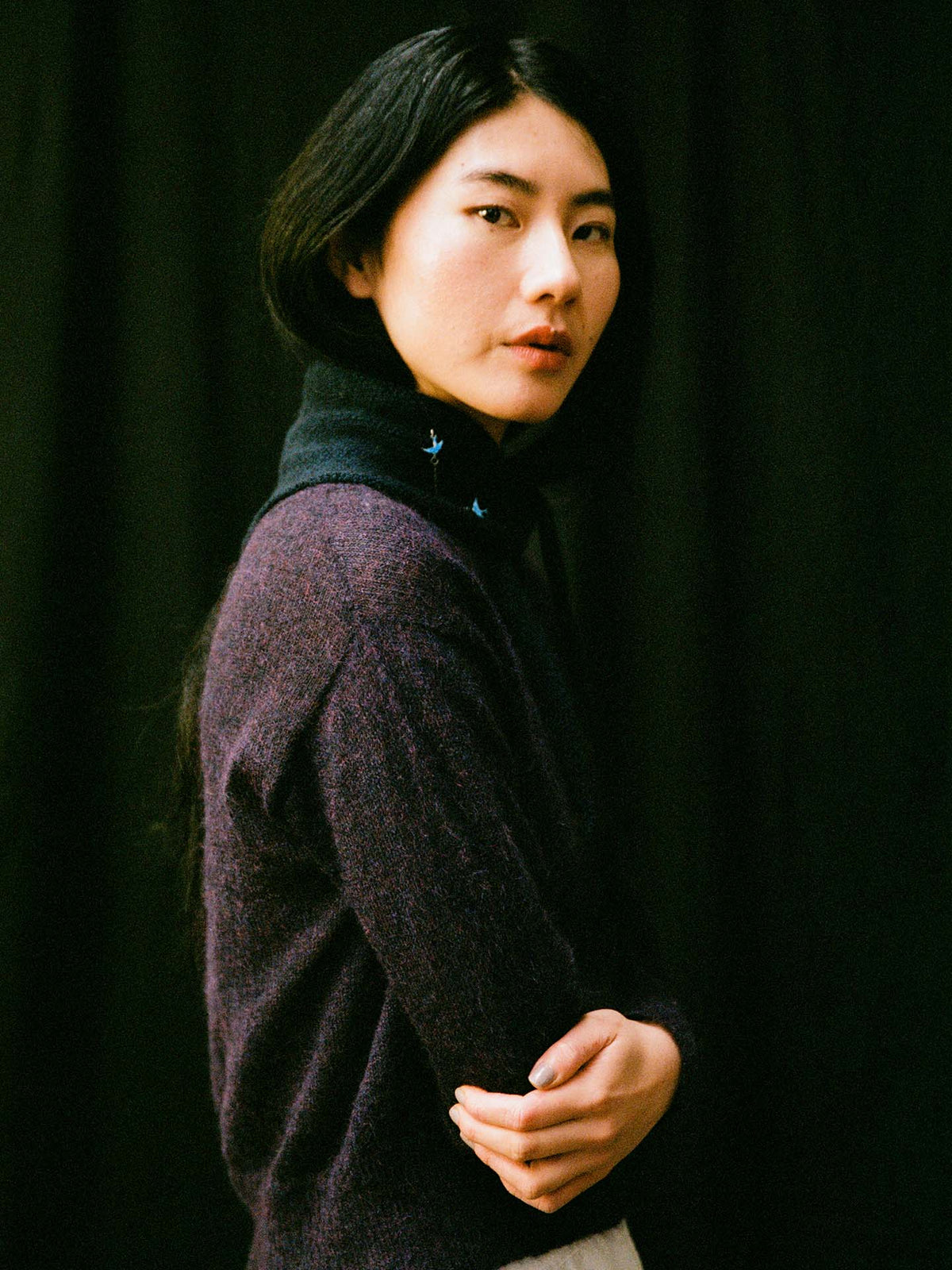 A person with long black hair, wearing a dark purple sweater and the Daisy Scarf – Ink (97cm x 22cm), stands against a black background, their composed look and neutral expression highlighted by the scarf.