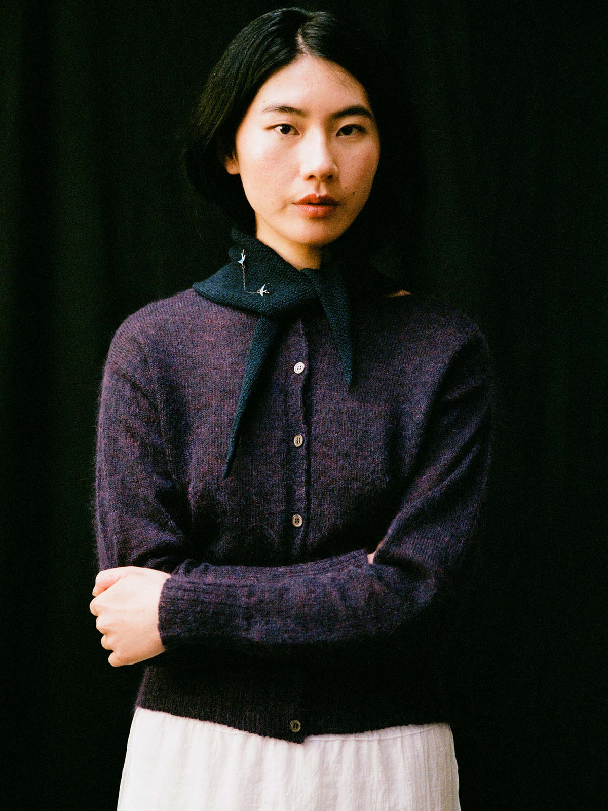 A person with straight black hair, wearing a purple button-up sweater, white skirt, and the Daisy Scarf – Ink (97cm x 22cm), stands against a black background with arms crossed.