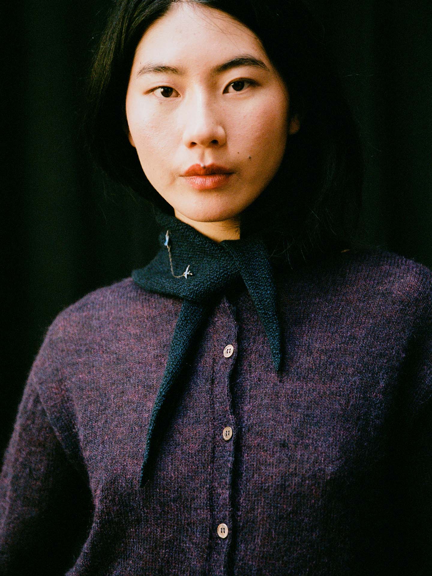 A person with long dark hair wears a purple button-up sweater and the Daisy Scarf – Ink, a dark scarf tied around their neck (22cm x 97cm), standing against a black background.