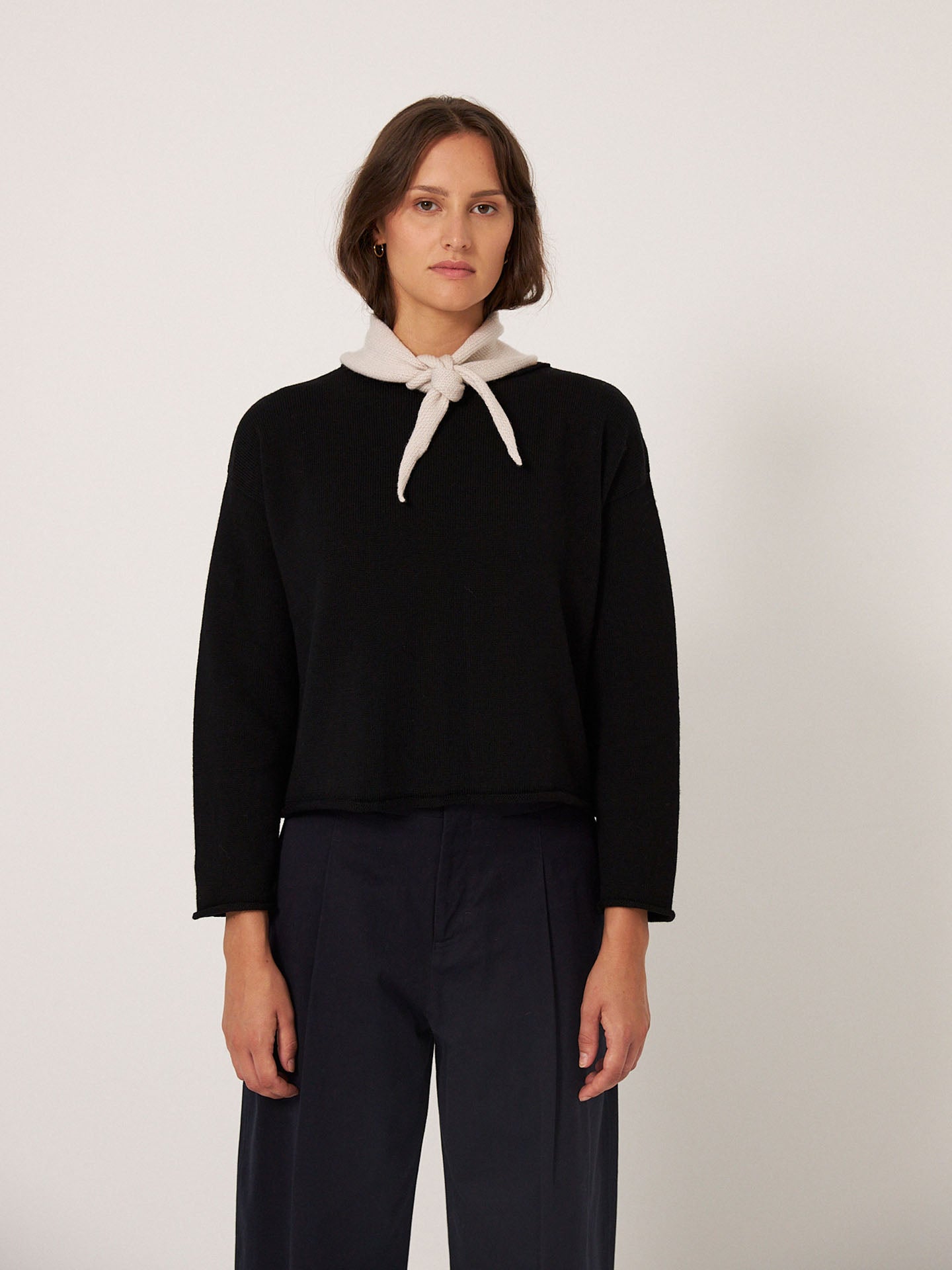 A person stands against a plain background wearing a black long-sleeve top, dark pants, and the Francie Daisy Scarf – Ivory (22cm wide, 97cm long) tied around their neck.