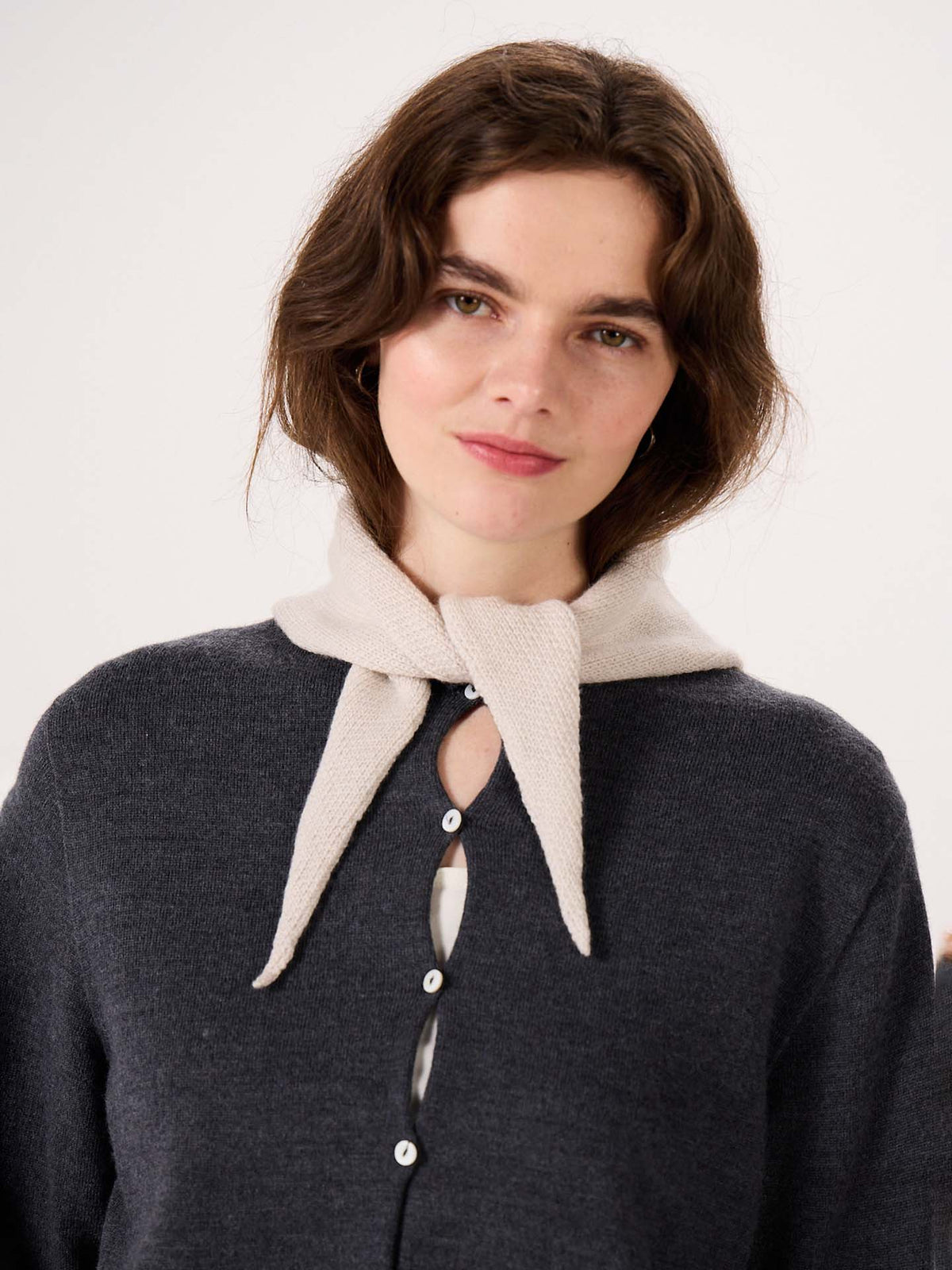 A woman with wavy brown hair stands against a plain background beside a wide fitness mat, wearing a dark buttoned cardigan and the Daisy Scarf in Ivory, tied loosely around her neck.