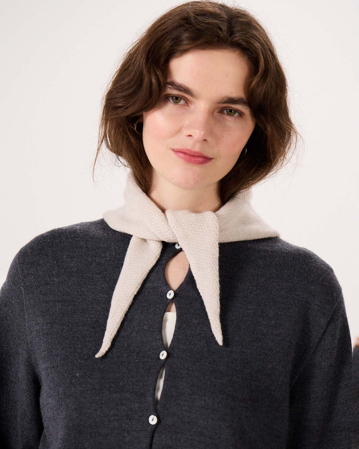A woman with wavy brown hair stands against a plain background beside a wide fitness mat, wearing a dark buttoned cardigan and the Daisy Scarf in Ivory, tied loosely around her neck.