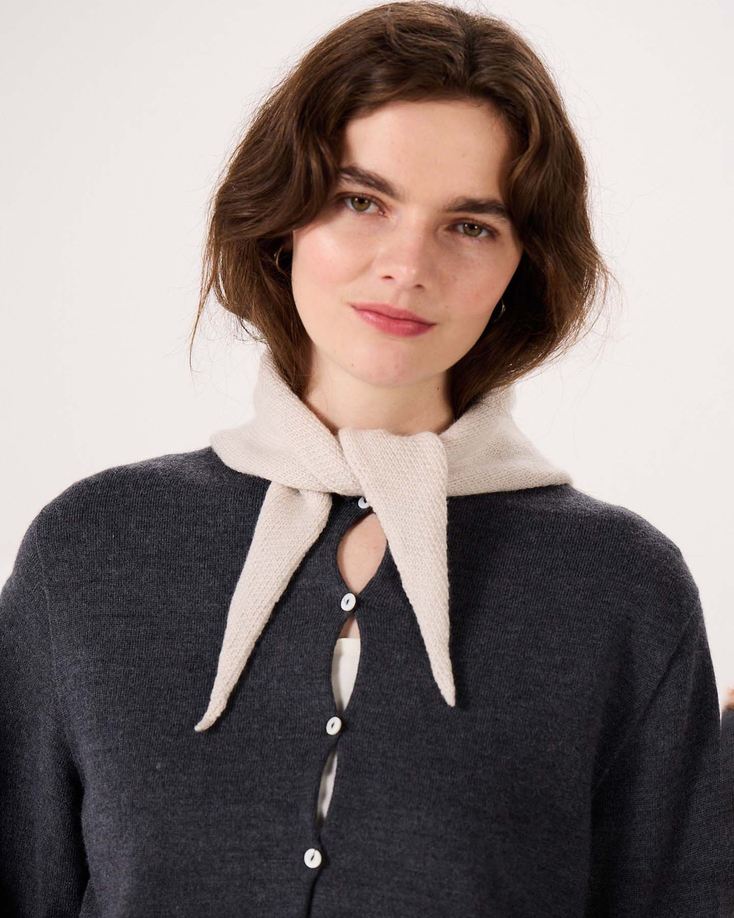 A woman with light skin and brown hair wears a dark gray buttoned sweater, with the Daisy Scarf in Ivory draped over her shoulders, standing next to a wide fitness mat against a plain light background.