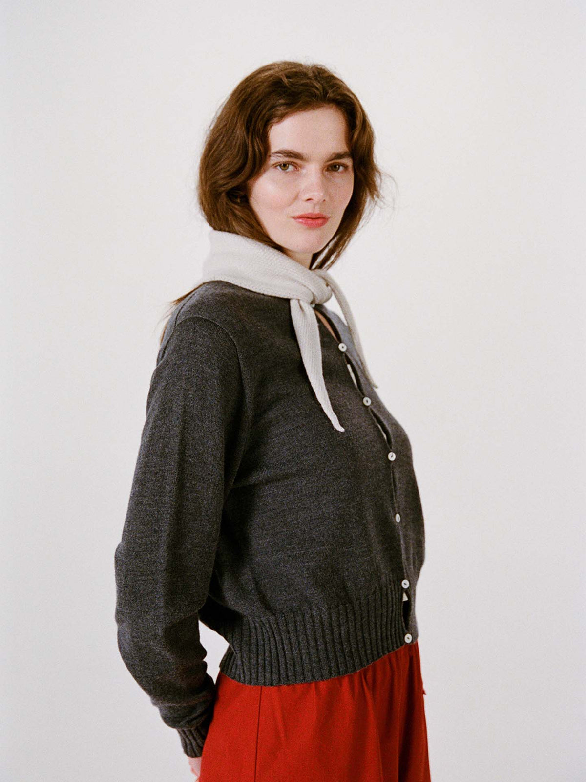 Woman wearing a dark gray buttoned sweater, bright red pants, and the Daisy Scarf in Ivory around her neck, standing against a plain white background on a wide fitness mat.
