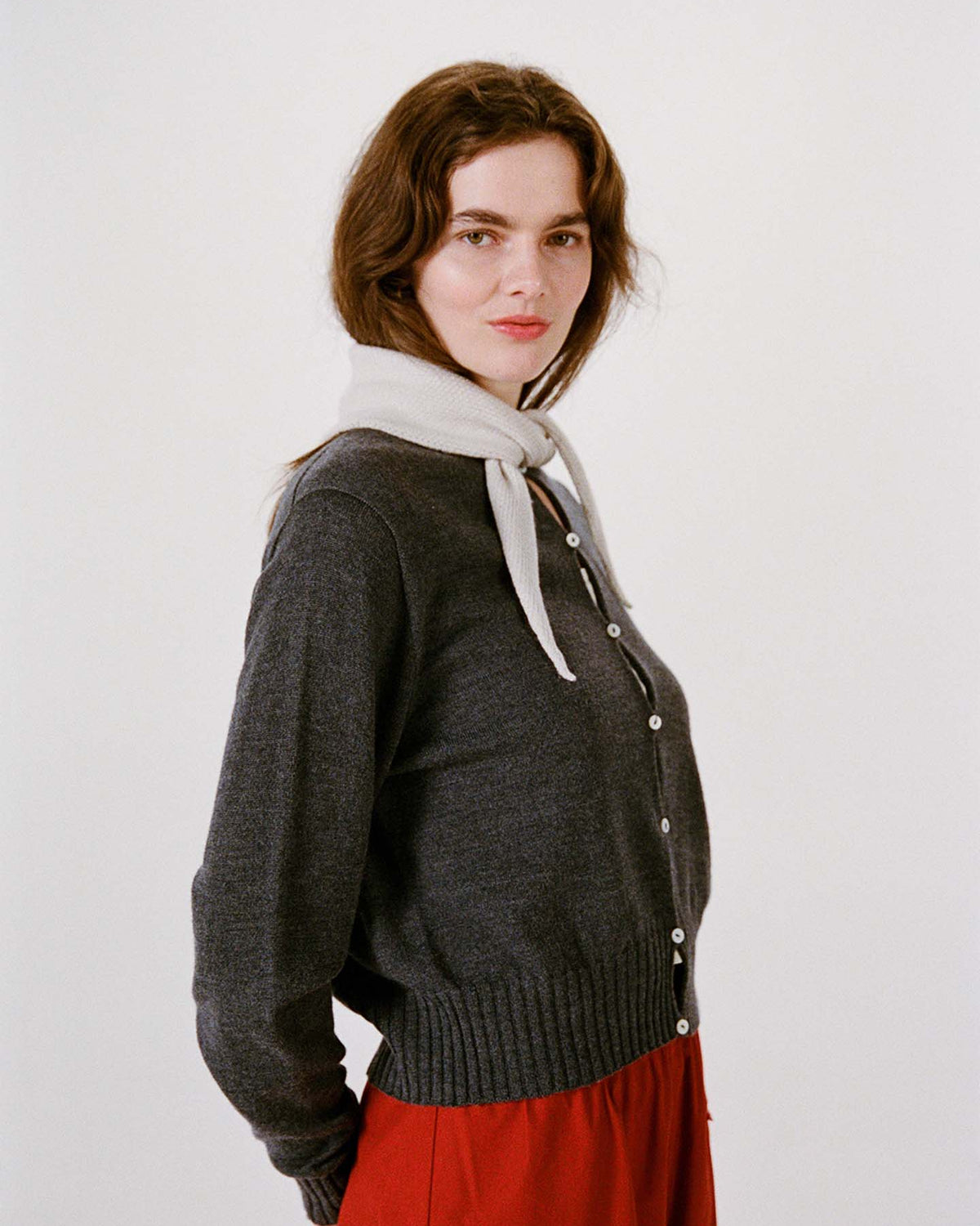Woman wearing a dark gray buttoned sweater, bright red pants, and the Daisy Scarf in Ivory around her neck, standing against a plain white background on a wide fitness mat.