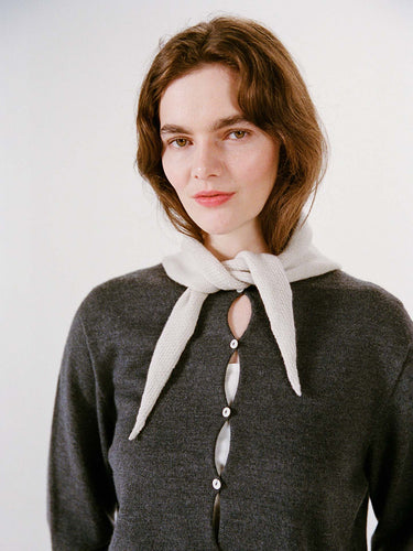 A woman with light skin and brown hair wears a dark gray buttoned sweater, with the Daisy Scarf in Ivory draped over her shoulders, standing next to a wide fitness mat against a plain light background.