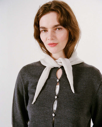 A woman with light skin and brown hair wears a dark gray buttoned sweater, with the Daisy Scarf in Ivory draped over her shoulders, standing next to a wide fitness mat against a plain light background.