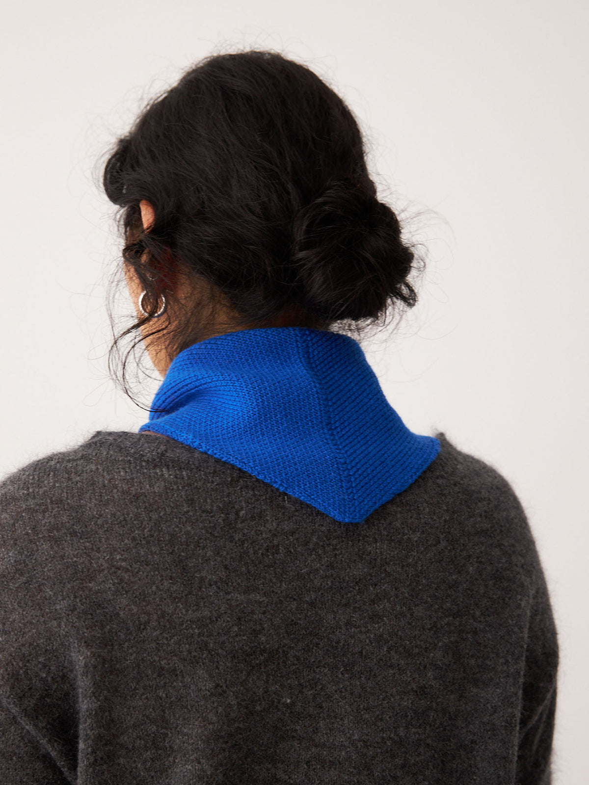 A person with dark hair in a low bun wears the Francie Daisy Scarf – Azure and a thick dark grey sweater, photographed from behind against a plain light background, standing next to an extra wide yoga mat.