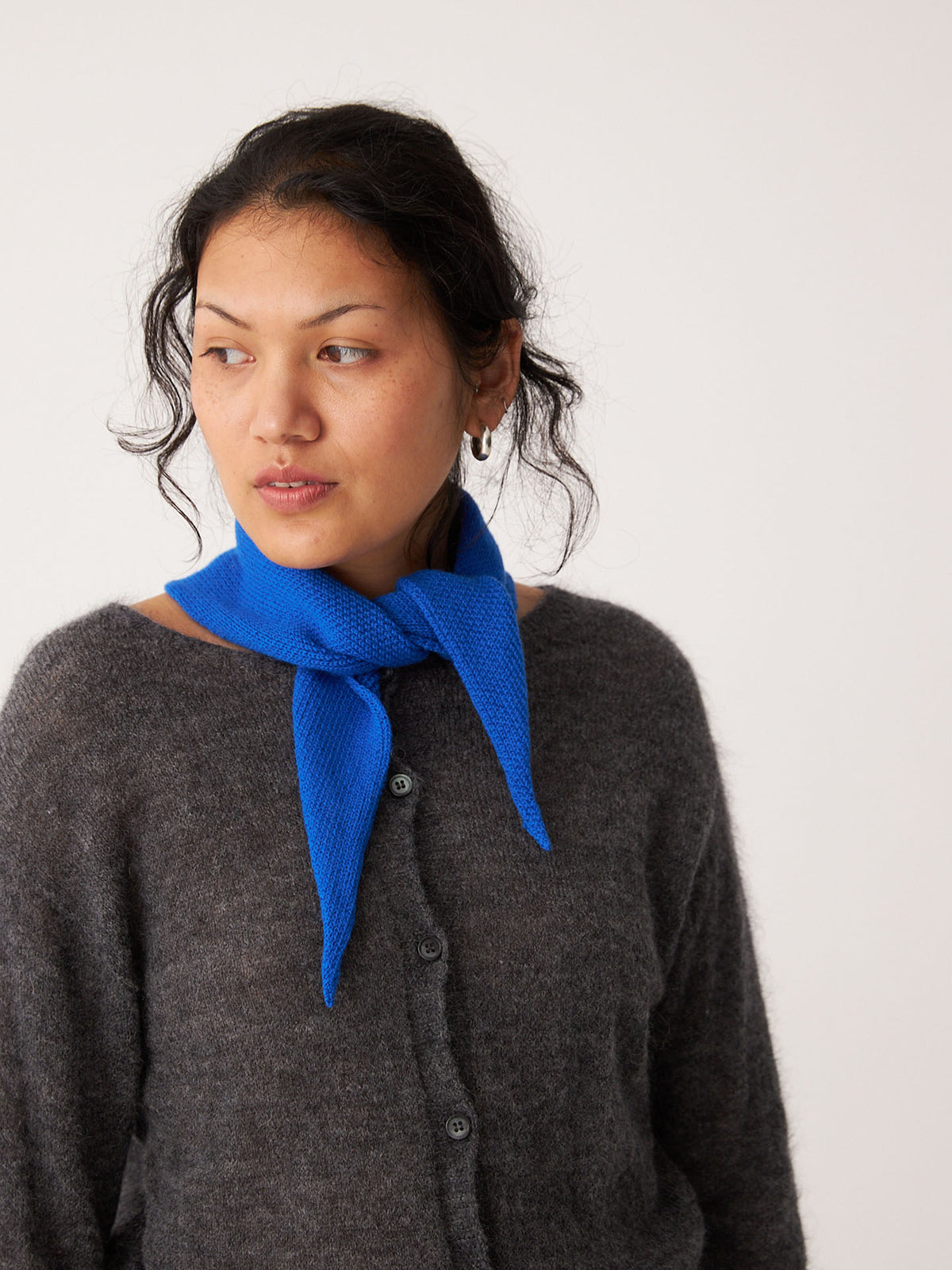 A person in a grey cardigan and Francie&#39;s Daisy Scarf – Azure stands next to an extra wide yoga mat against a plain light background, looking to the side.