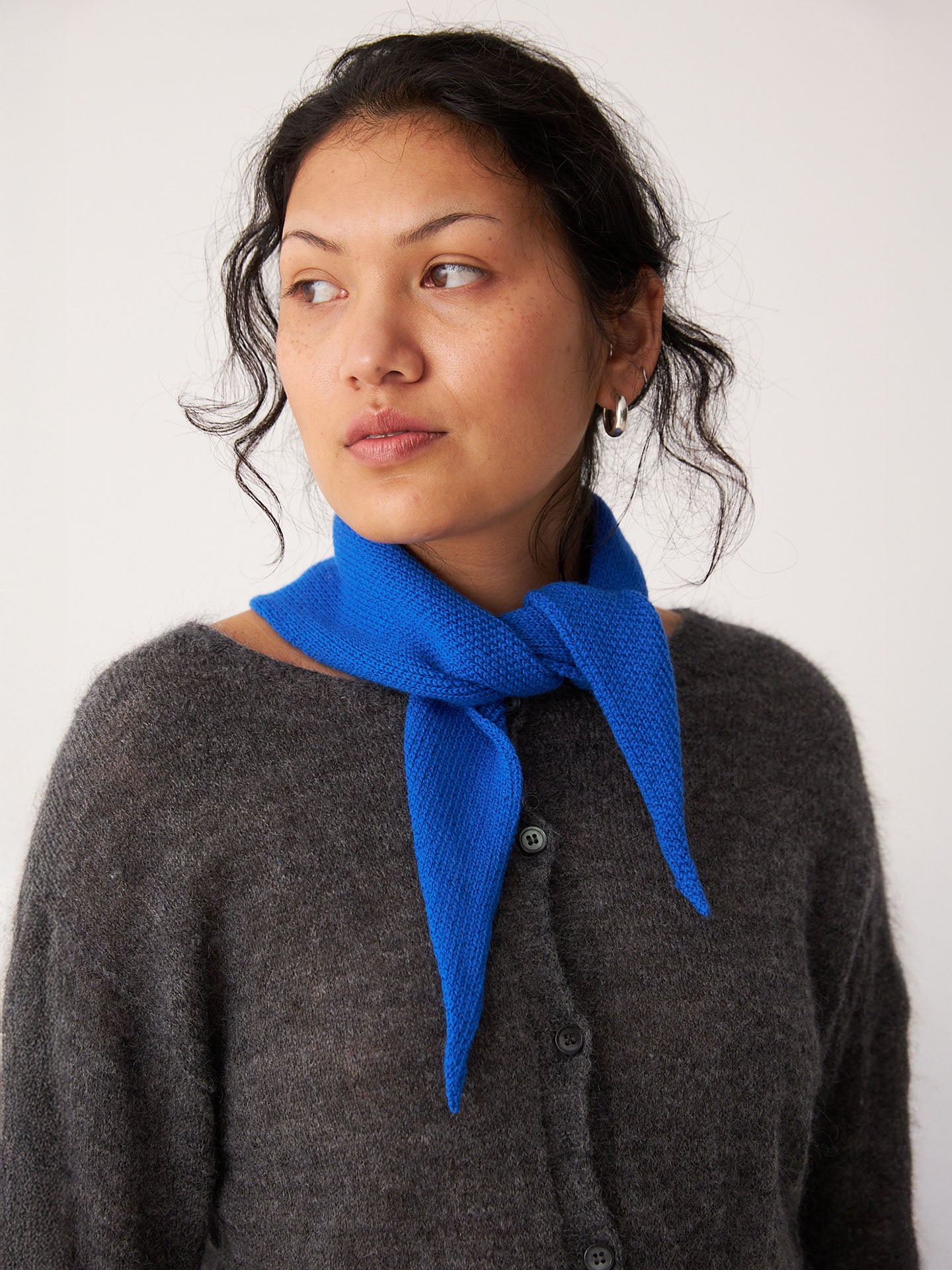 A person wearing a grey cardigan and the Francie Daisy Scarf – Azure, 97cm by 22cm, tied around their neck, looking slightly to the side against a plain background.