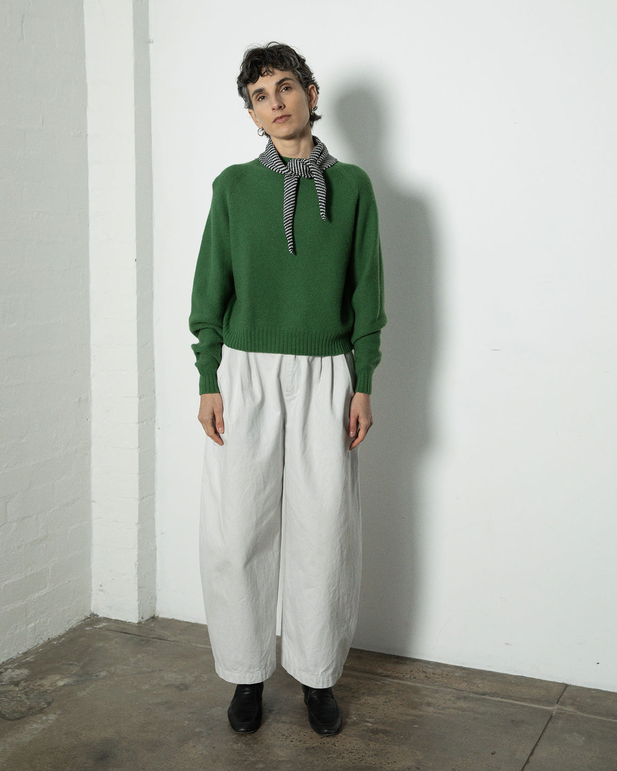 A person stands indoors against a white wall, wearing a green sweater, white wide-leg pants, black shoes, and the Daisy Scarf – Black & Creme Stripe (97cm x 22cm).