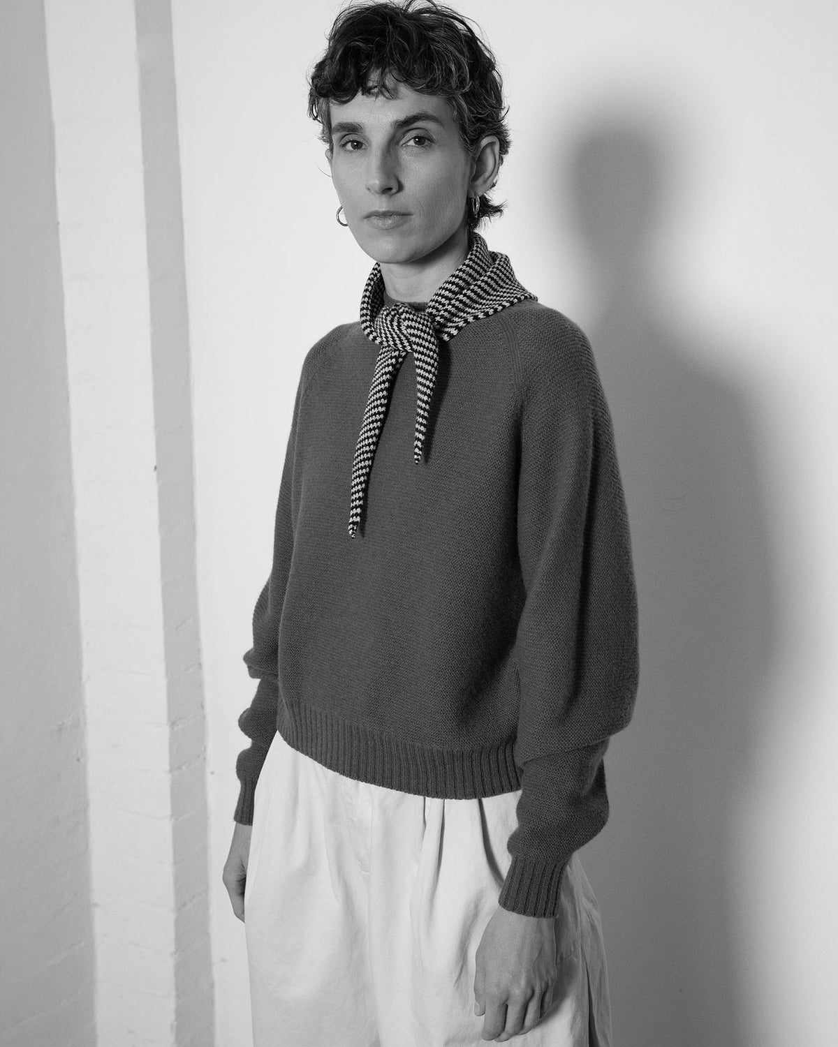 A person with short curly hair wears a sweater, wide pants, and the Daisy Scarf – Black & Creme Stripe (97cm x 22cm) tied around their neck, standing against a plain wall in a black and white photo.