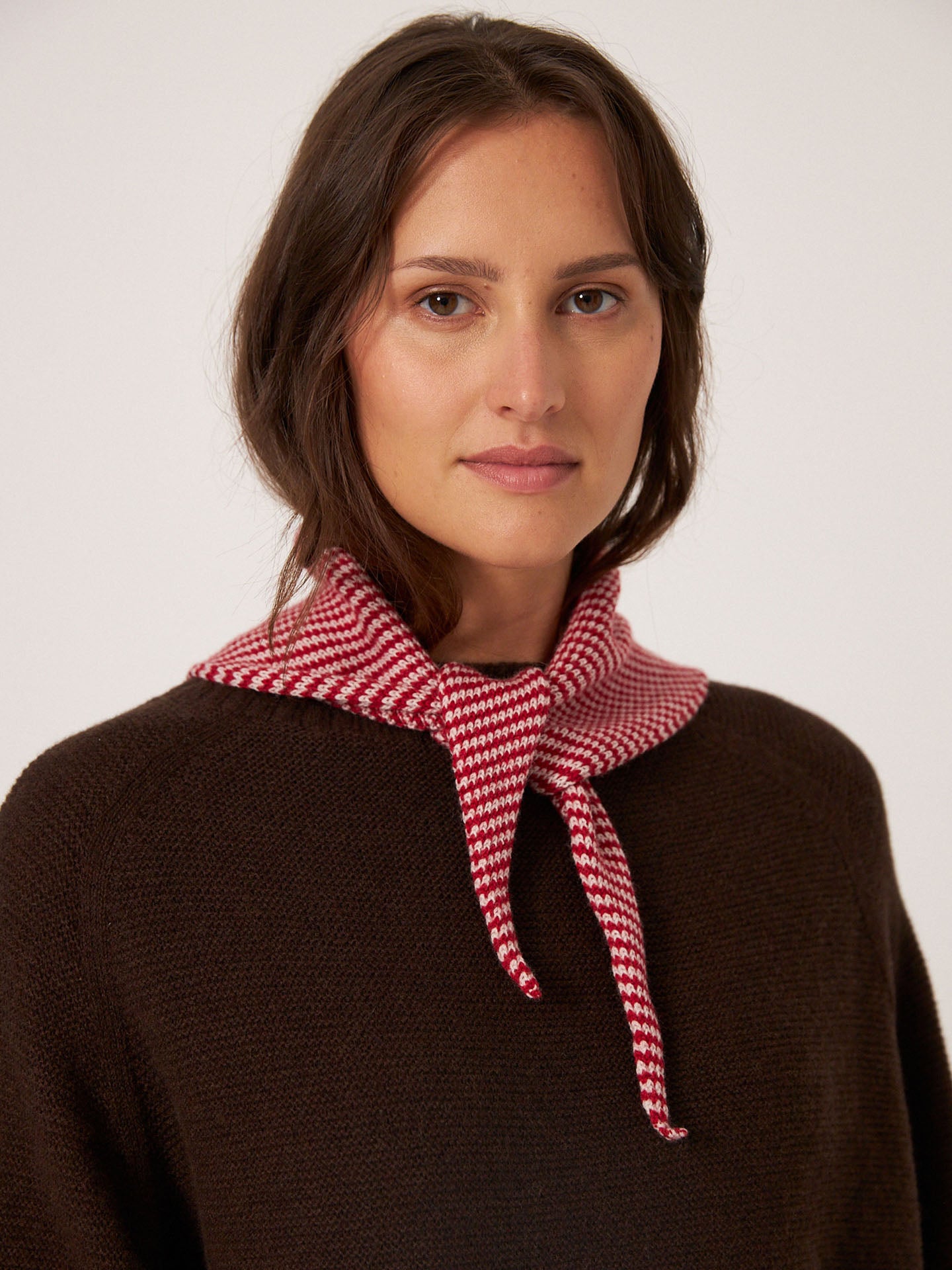 A woman stylishly drapes the Francie Daisy Scarf – Cherry Stripe over her brown top, with the knot highlighting its 22cm by 97cm dimensions against a plain background.