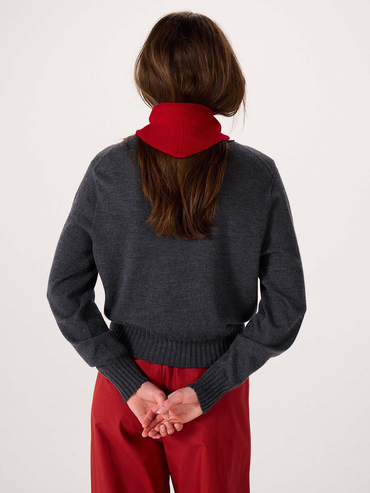 A person with long brown hair stands facing away, wearing a dark gray sweater, red pants, and the Daisy Scarf – Tamarillo. The scarf’s bold silhouette is accentuated by its dimensions of 97cm long and 22cm wide, as the person clasps their hands behind their back against a plain background.
