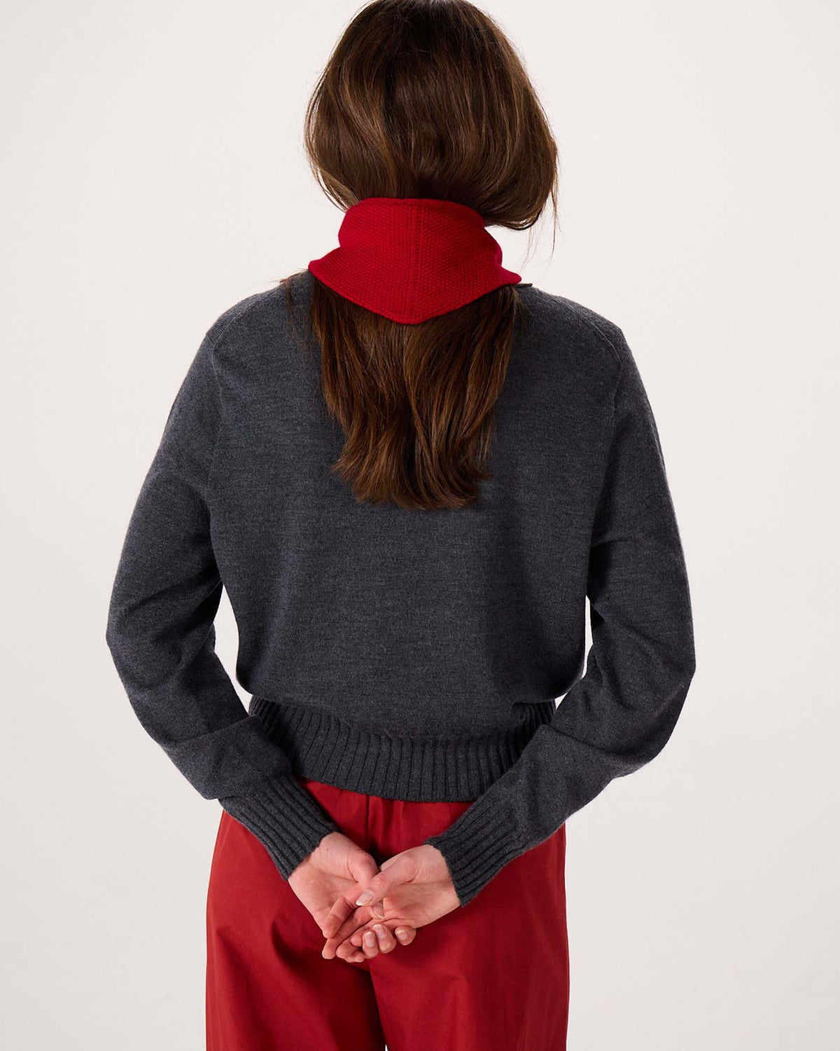 A person with long brown hair stands facing away, wearing a dark gray sweater, red pants, and the Daisy Scarf – Tamarillo. The scarf’s bold silhouette is accentuated by its dimensions of 97cm long and 22cm wide, as the person clasps their hands behind their back against a plain background.