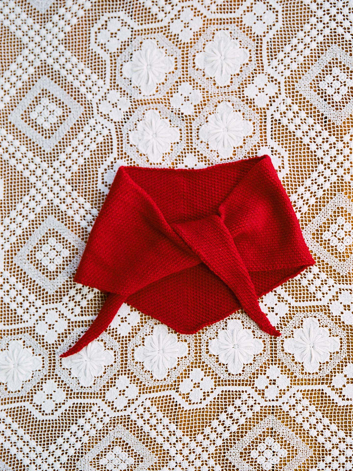 The Daisy Scarf – Tamarillo, a red knitted scarf with pointed ends measuring 22cm wide and 97cm long, is laid flat on a white lace tablecloth featuring a geometric floral pattern.