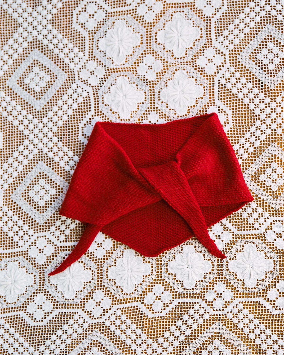The Daisy Scarf – Tamarillo, a red knitted scarf with pointed ends measuring 22cm wide and 97cm long, is laid flat on a white lace tablecloth featuring a geometric floral pattern.