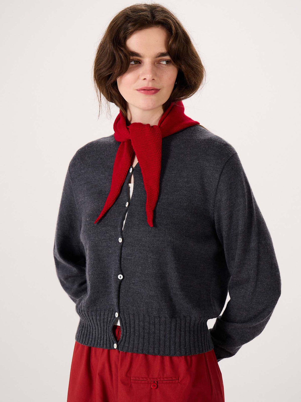 A woman stands against a plain light background, wearing a dark gray button-up cardigan and red pants, her look complemented by the Daisy Scarf – Tamarillo stylishly tied around her neck. Measuring 97cm long and 22cm wide, this scarf makes the perfect accessory to complete her outfit.