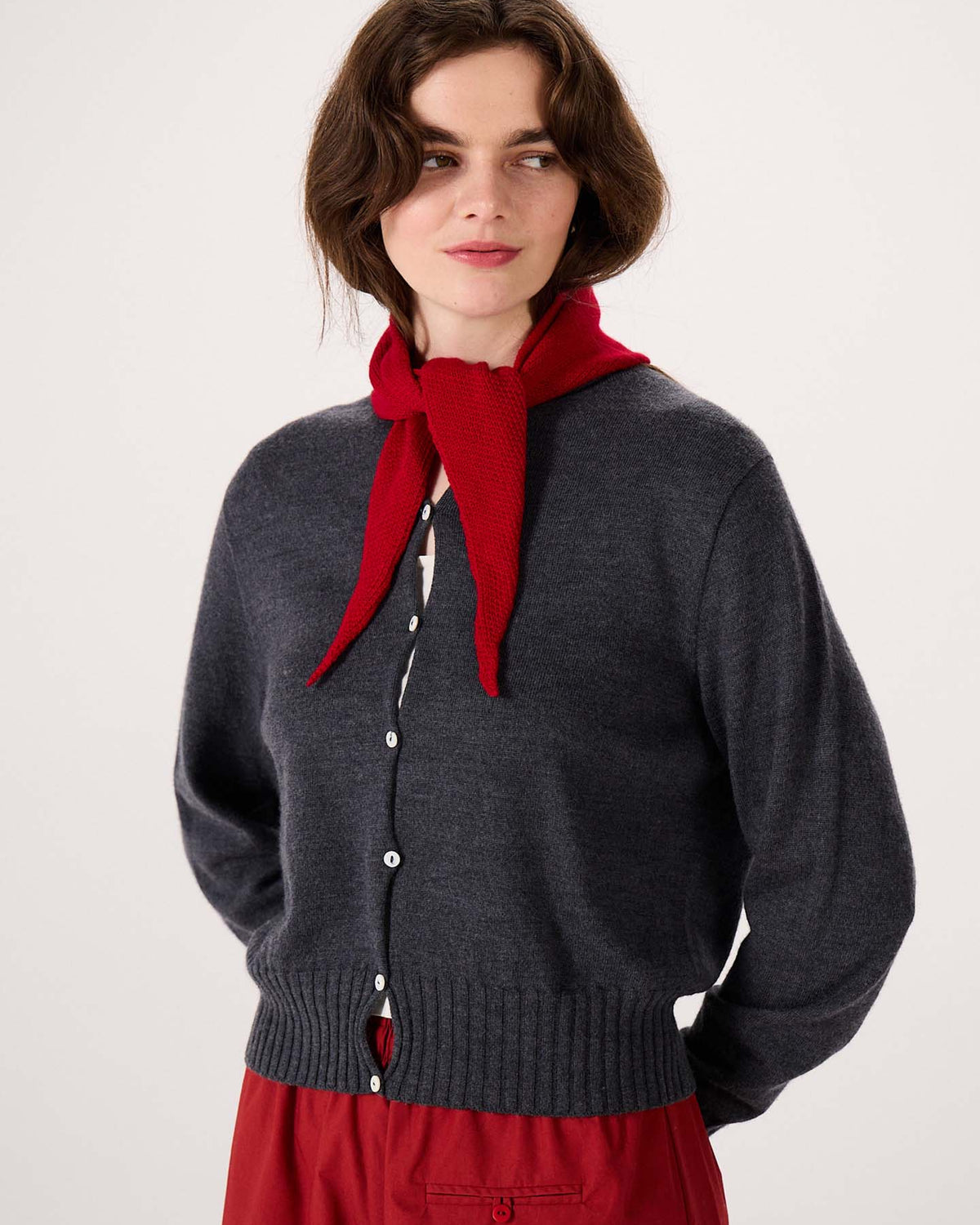 A woman stands against a plain light background, wearing a dark gray button-up cardigan and red pants, her look complemented by the Daisy Scarf – Tamarillo stylishly tied around her neck. Measuring 97cm long and 22cm wide, this scarf makes the perfect accessory to complete her outfit.