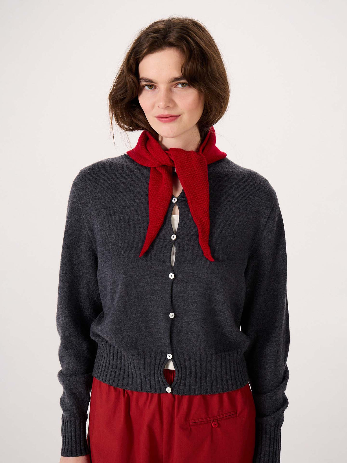 A woman stands against a plain white background wearing a dark grey button-up cardigan, a red skirt, and the Daisy Scarf – Tamarillo tied around her neck. The scarf measures 22cm wide by 97cm long, providing stylish and comfortable coverage.