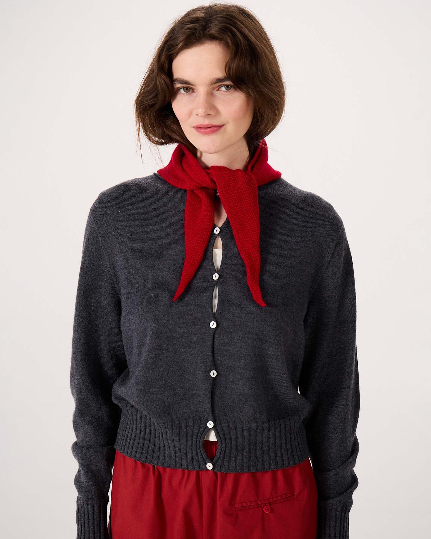 A woman with wavy brown hair is wearing a dark gray buttoned sweater and the Daisy Scarf – Tamarillo, tied around her neck, standing against a plain light background. The scarf measures 97cm long and 22cm wide, offering stylish comfort with practical product dimensions.